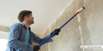 Mann mit Schutzbrille trägt mit langem Stiel und Rolle Öko Tiefengrund auf eine rohe Wand auf.