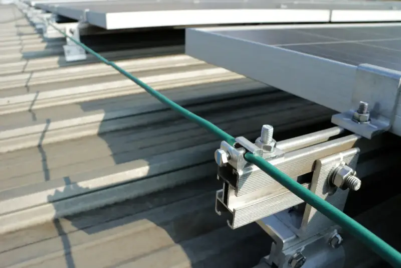 Detailaufnahme der Erdung einer PV-Anlage. Ein grünes Kabel wird sicher mit einer Klemme an der Aluminiumkonstruktion befestigt, die die Solarmodule auf dem Dach hält.